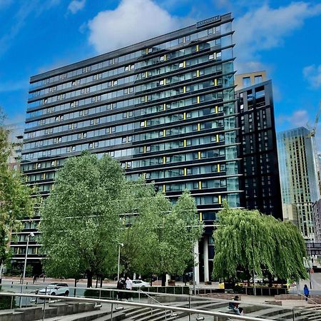 Citysuites Aparthotel Salford  Exterior photo The photo shows a modern multi-story building with a glass and steel facade, surrounded by trees and greenery. The structure features a series of horizontal lines and large windows, reflecting the sky and nearby buildings. In the foreground, there ar