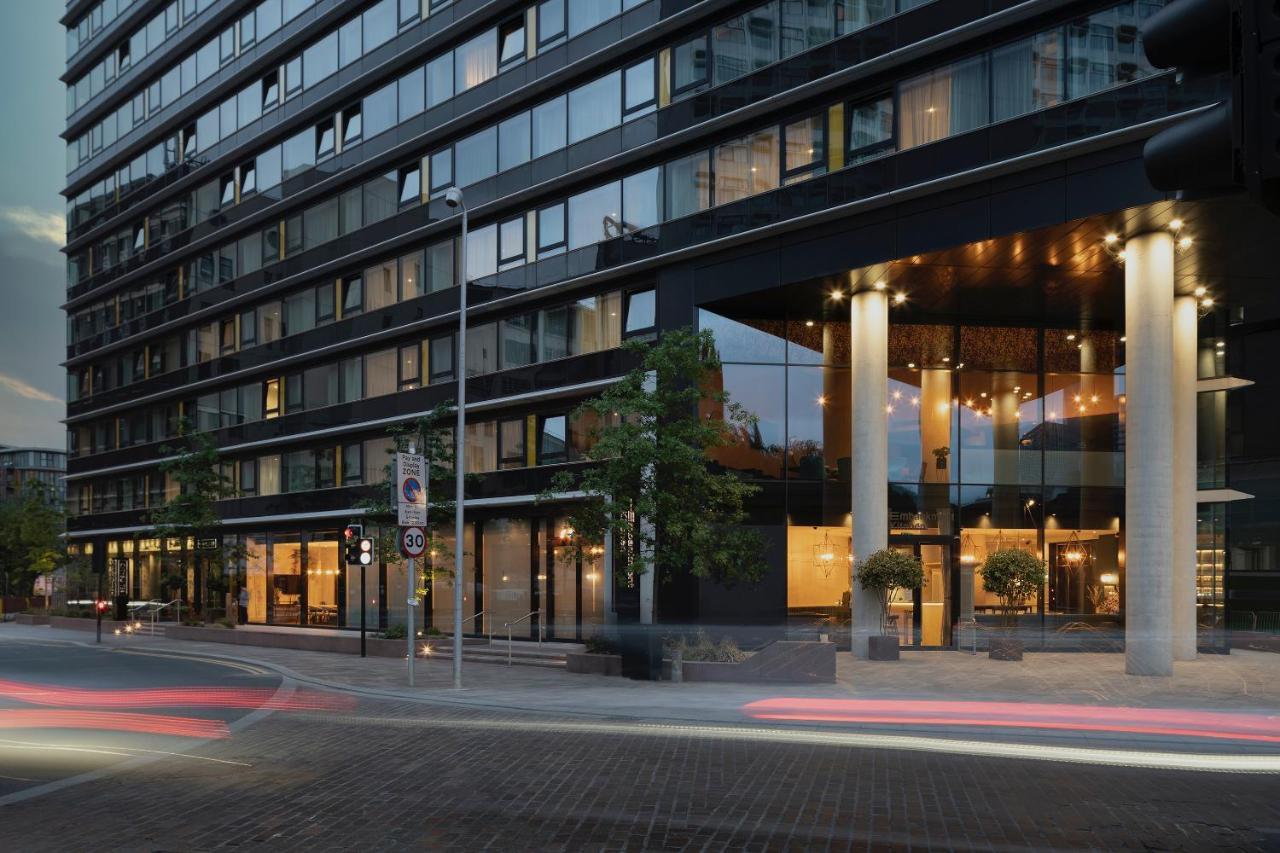 Citysuites Aparthotel Salford  Exterior photo The photo shows a modern multi-story building with a sleek glass facade. The ground level features a well-lit entrance, characterized by tall pillars and decorative lighting, suggesting it may be a hotel or a stylish commercial space. There are trees