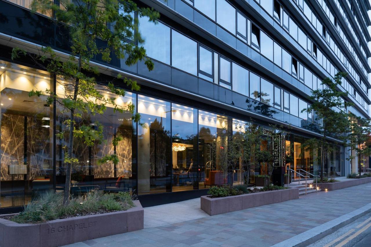 Citysuites Aparthotel Salford  Exterior photo The photo shows a modern building with a glass facade, reflecting the surrounding environment. The structure has several floors and features large windows that allow visibility into the interior, which appears well-lit. There are trees and greenery p