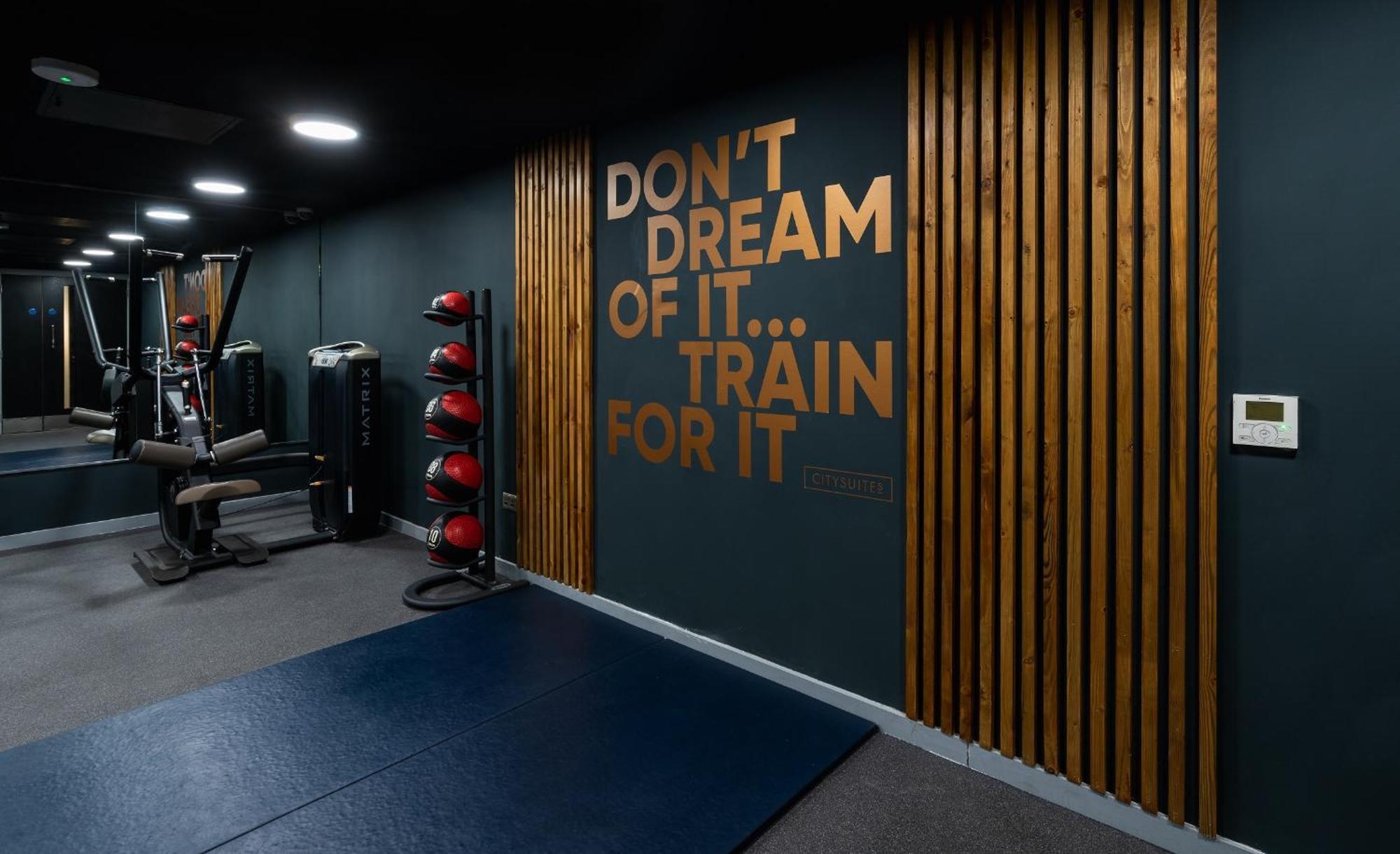 Citysuites Aparthotel Salford  Exterior photo The photo depicts a section of a gym or fitness area. The wall features a motivational quote that reads: "DON'T DREAM OF IT... TRAIN FOR IT," highlighted in large, bold letters. The wall is designed with a combination of dark green paint and wooden s