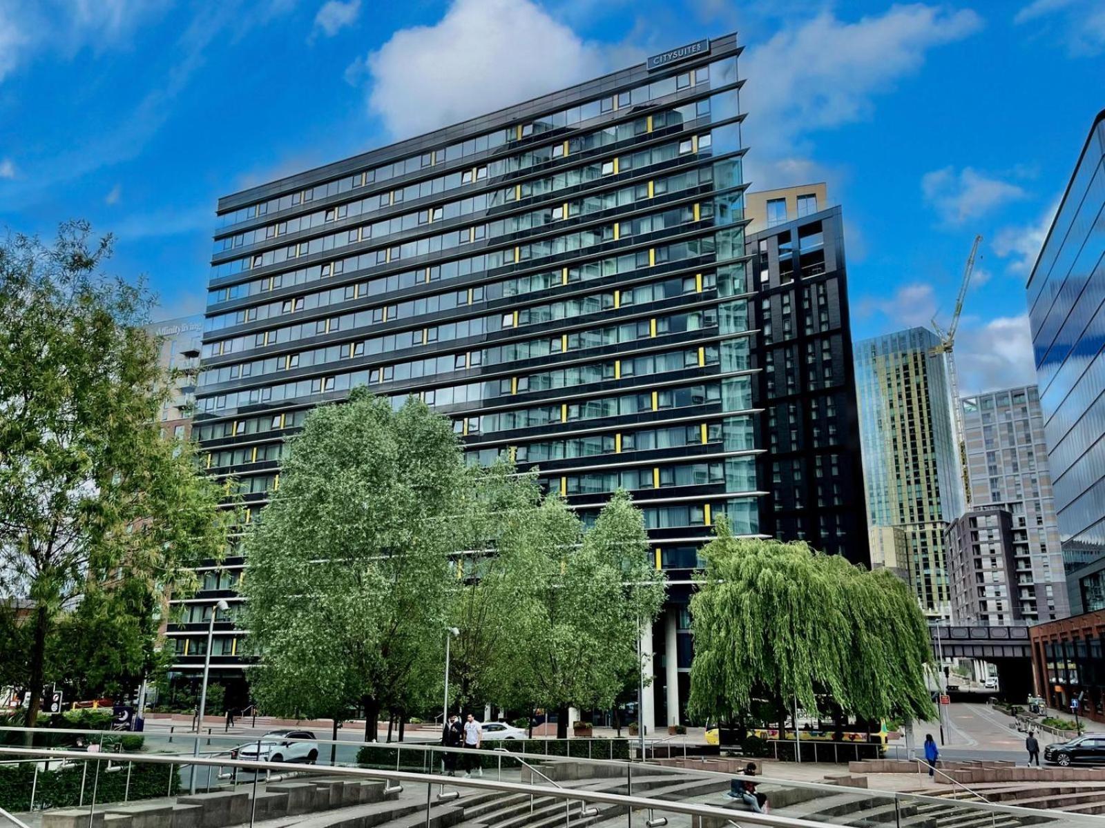 Citysuites Aparthotel Salford  Exterior photo The photo shows a modern multi-story building with a glass and steel facade, surrounded by trees and greenery. The structure features a series of horizontal lines and large windows, reflecting the sky and nearby buildings. In the foreground, there ar
