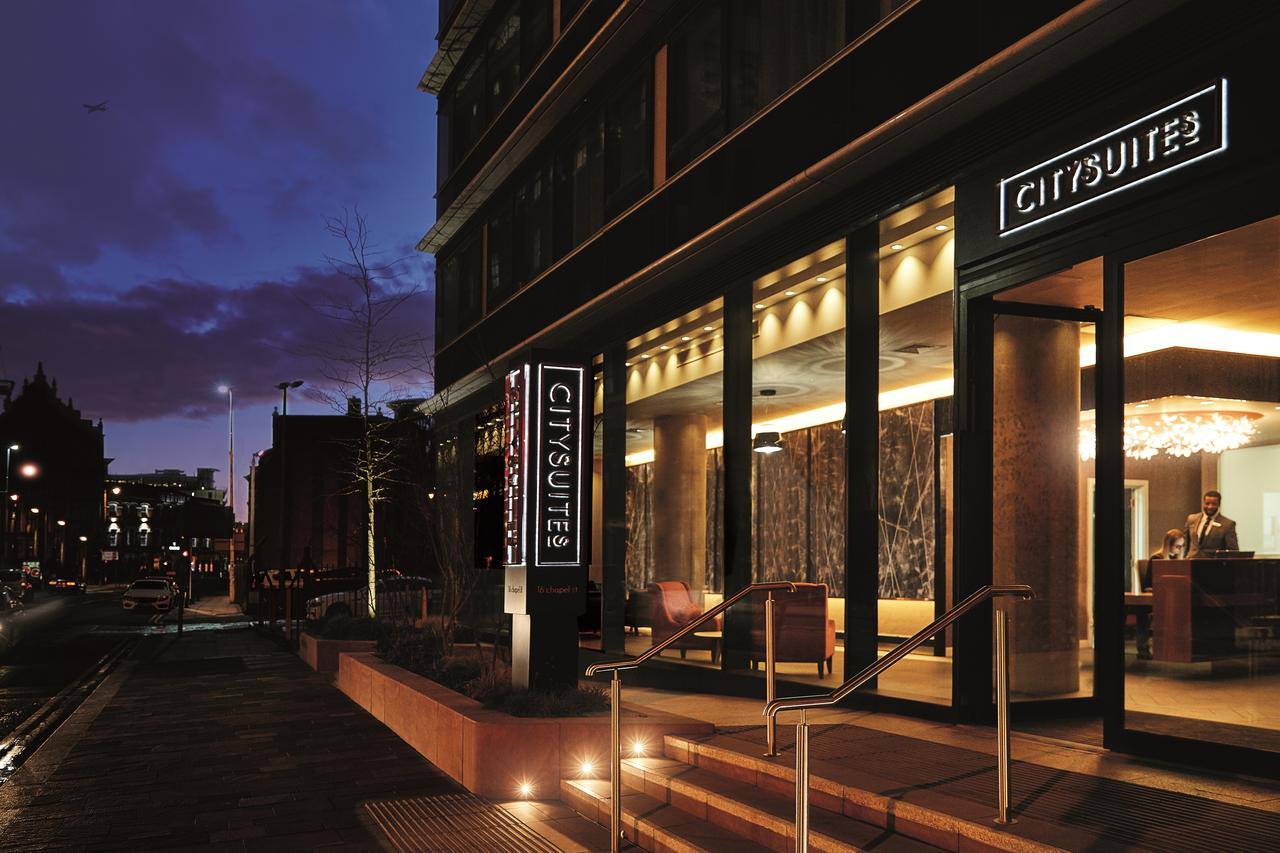 Citysuites Aparthotel Salford  Exterior photo The photo shows the exterior of a modern hotel named "CitySuites," taken at dusk. The entrance features large glass doors and an illuminated sign displaying the hotel's name. The building has a contemporary design with a sleek facade and warm lightin