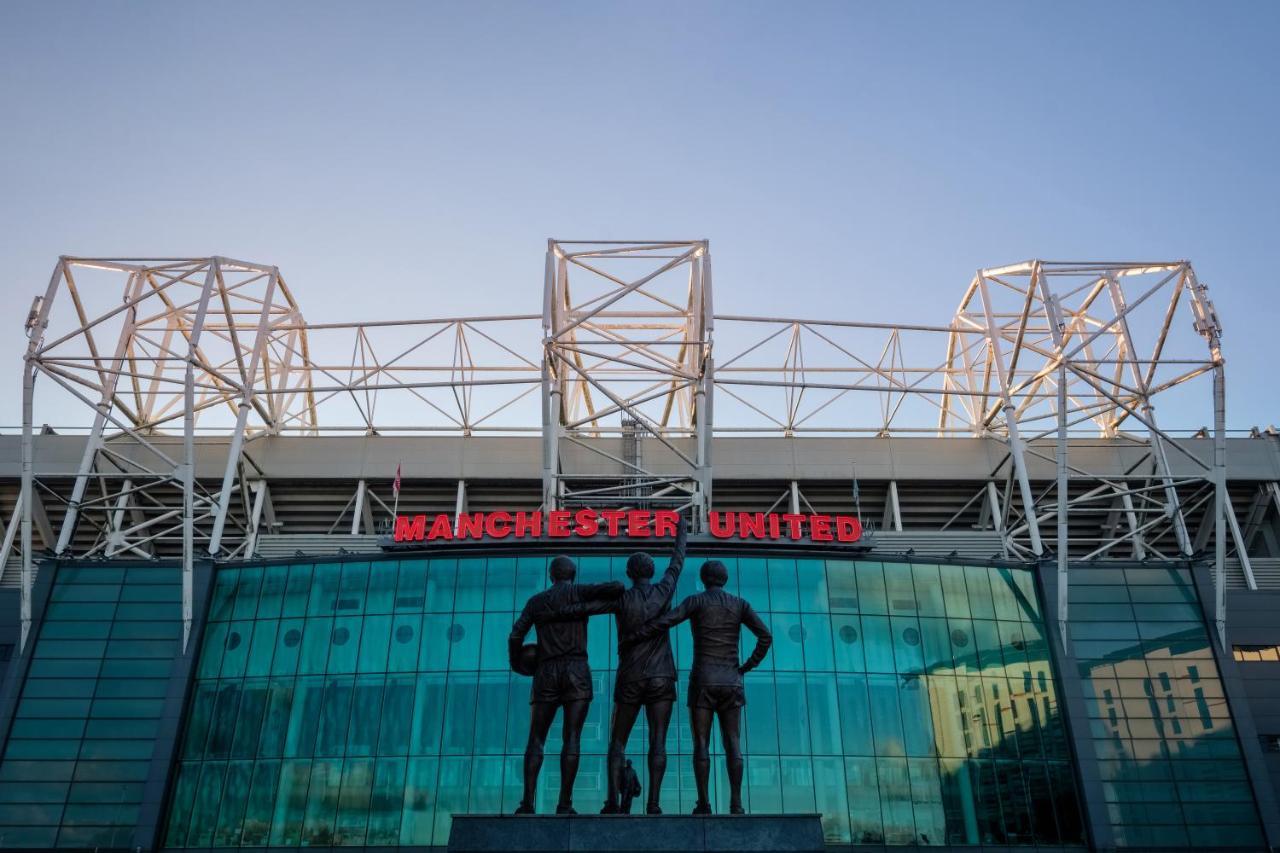 Citysuites Aparthotel Salford  Exterior photo The photo shows the exterior of Old Trafford, the home stadium of Manchester United Football Club. In front of the stadium, there is a statue depicting three iconic figures from the club's history, known as the "United Trinity." The stadium has a mod