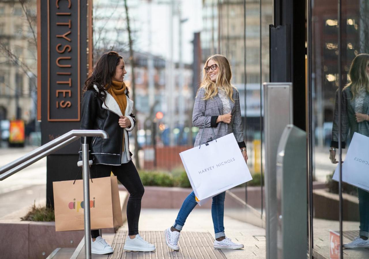Citysuites Aparthotel Salford  Exterior photo The photo shows two women walking together near a building entrance. One woman is wearing a black jacket with a hood, black pants, and white sneakers, while holding a large bag from a store. The other woman has long blonde hair and is wearing a gray 