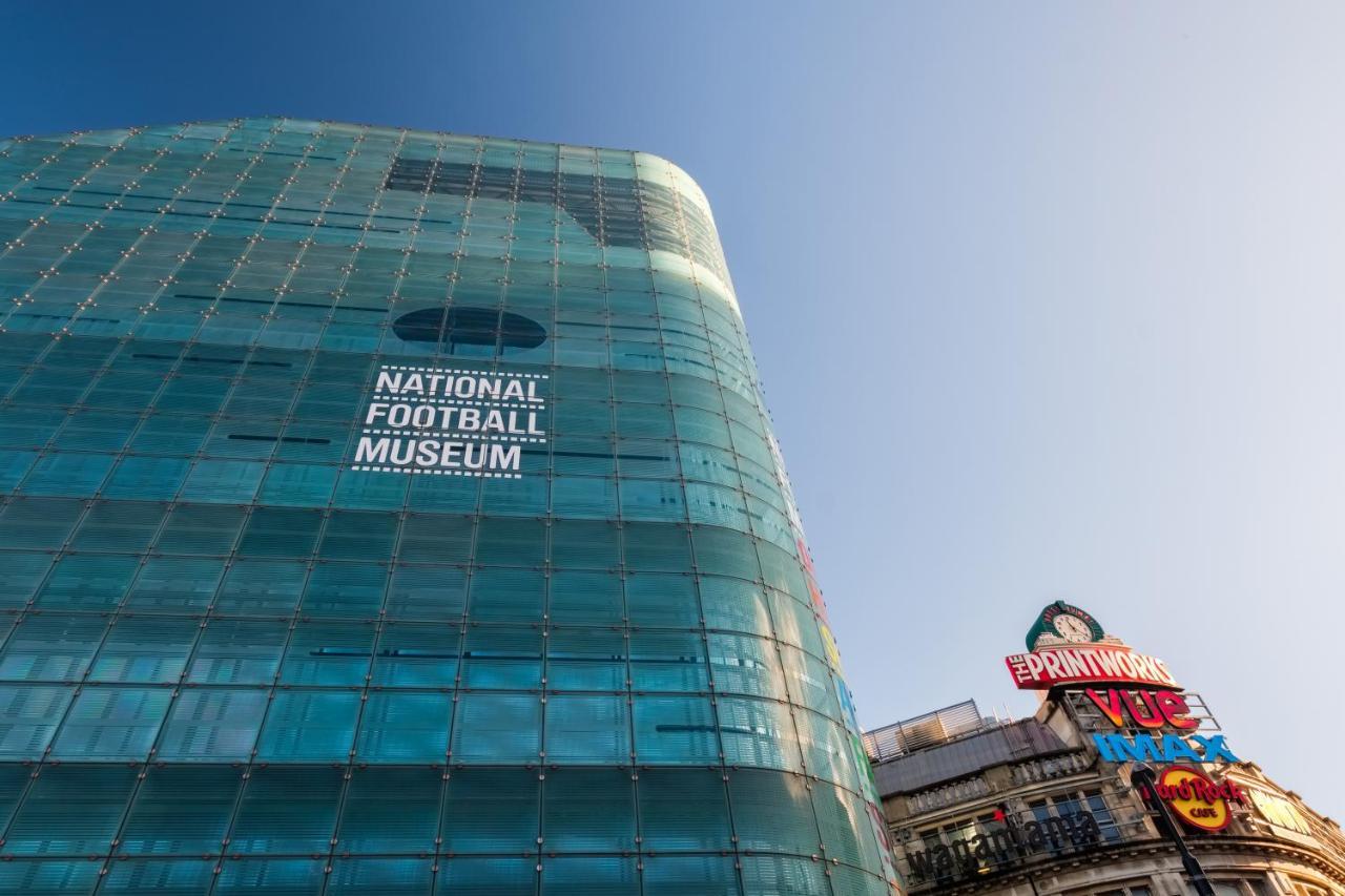 Citysuites Aparthotel Salford  Exterior photo The photo shows a modern glass building with the words "NATIONAL FOOTBALL MUSEUM" prominently displayed on one side. The building has a sleek, contemporary design, reflecting light in various shades. In the foreground, there is a colorful sign on a n