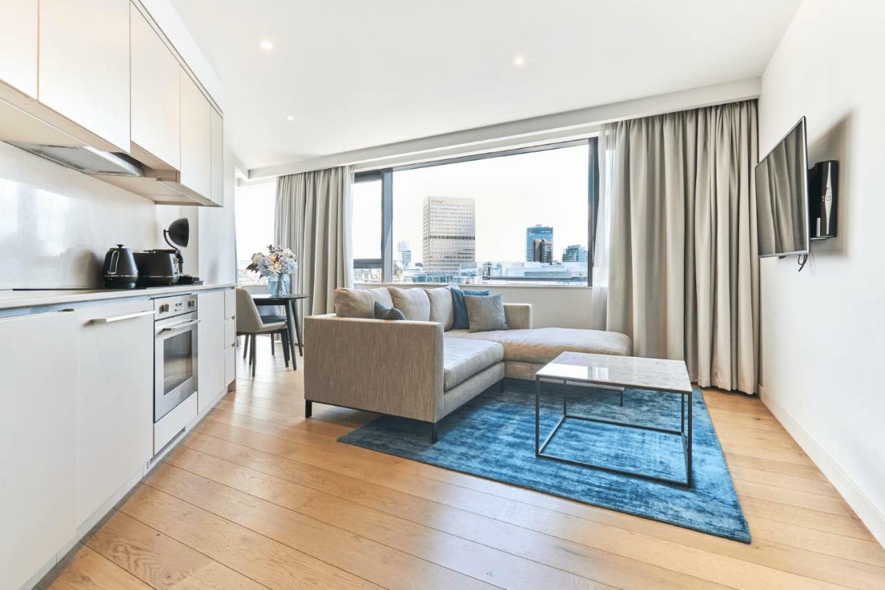 Citysuites Aparthotel Salford  Exterior photo The photo shows a modern living space with a stylish design. On one side, there is a kitchenette featuring a stove, oven, kettle, and other appliances, all in a light color. The main living area includes a light gray sofa positioned on a blue area ru