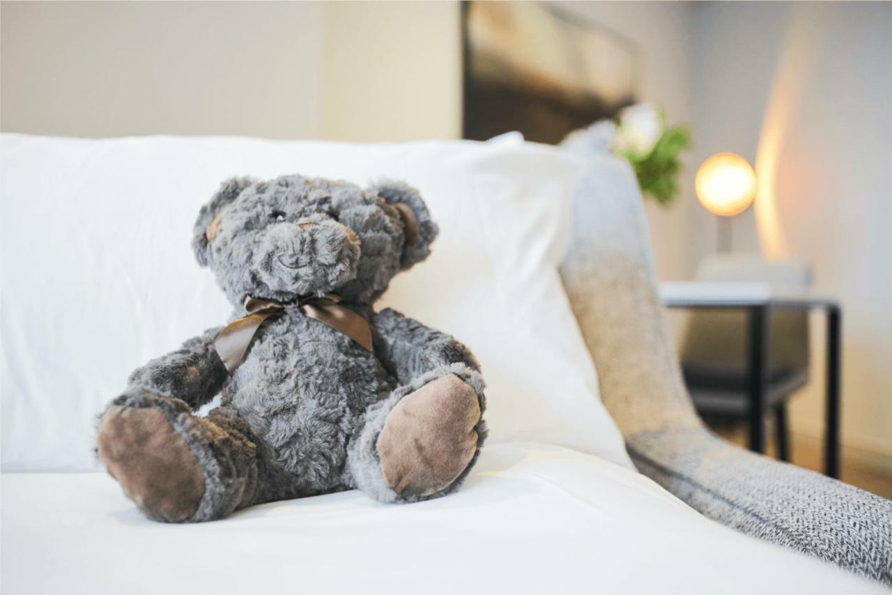 Citysuites Aparthotel Salford  Exterior photo The photo shows a plush teddy bear, which is gray in color, sitting on a white couch. The bear has a brown ribbon tied around its neck and has seen some wear, with noticeable shading on its paws. In the background, there is a softly lit environment w