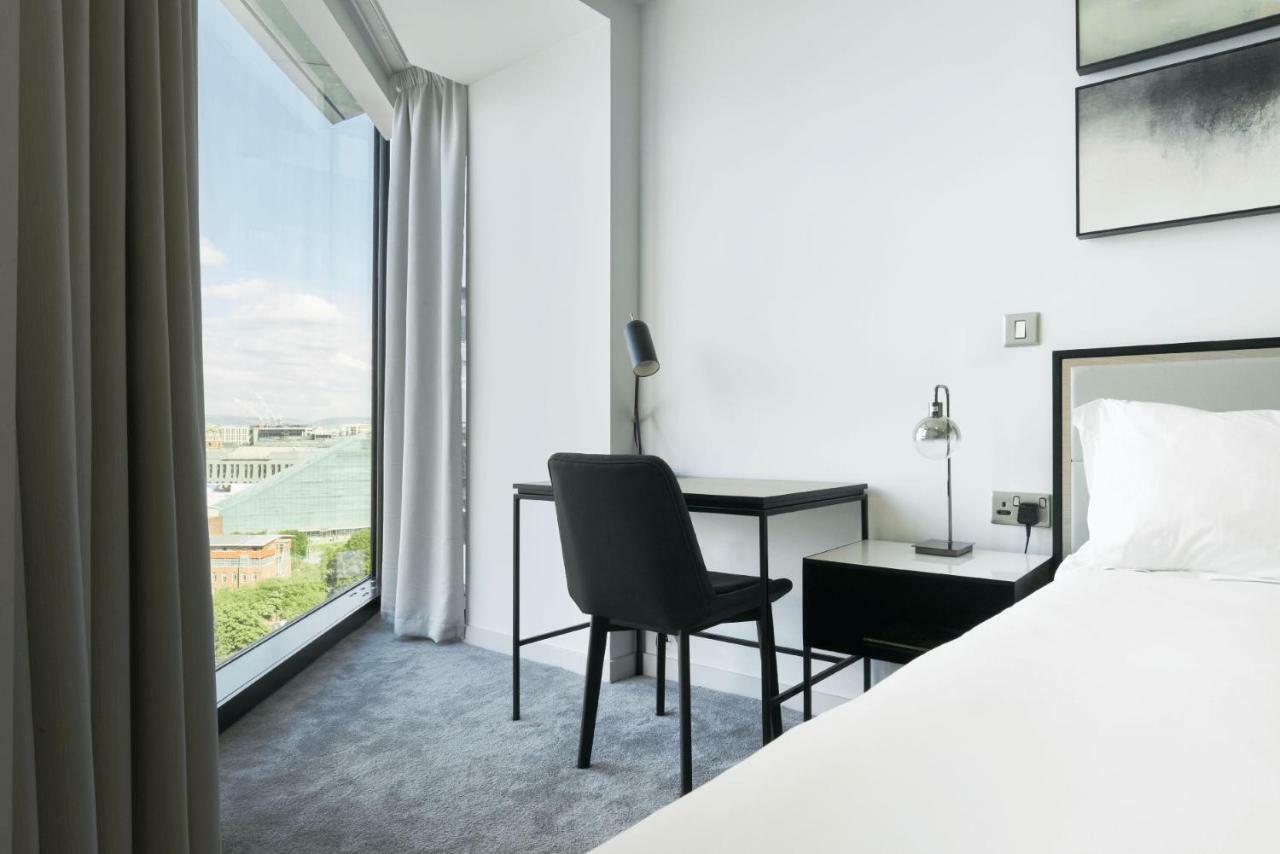 Citysuites Aparthotel Salford  Exterior photo The photo shows a modern hotel room featuring a bed with crisp white linens. To one side, there is a large window with a view of the outside, framed by sheer curtains that allow natural light to enter. A sleek black desk and chair are positioned next